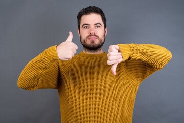 Young handsome Caucasian man wearing yellow sweater against gray wall showing thumbs up and thumbs down, difficult choose concept - obrazy, fototapety, plakaty