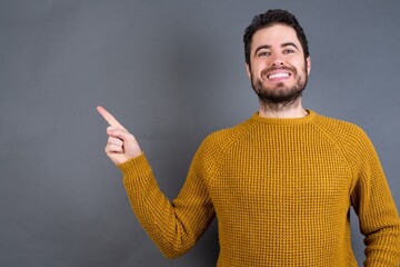 Fototapeta premium Young handsome Caucasian man wearing yellow sweater against gray wall looking at camera indicating finger empty space sales