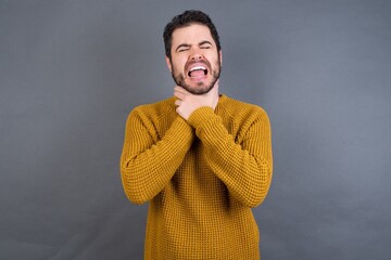 Young handsome Caucasian man wearing yellow sweater against gray wall shouting suffocate because...