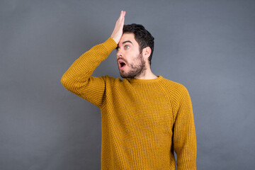 Young handsome Caucasian man wearing yellow sweater against gray wall surprised with hand on head...