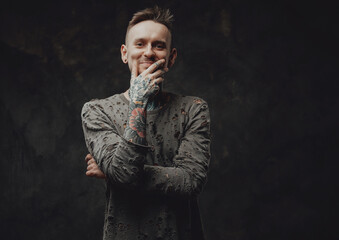 Portraif of a happy and tattooed hipster person dressed in ragged gray shirt posing in dark background with hand under his chin.