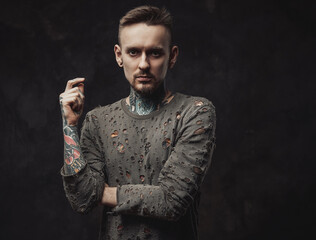 Portrait of a bearded and fashionable male hipster with tattoos and modern in gray ragged shirt hairstyle posing in dark background.