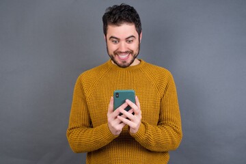 Young handsome Caucasian man wearing yellow sweater against gray wall using mobile phone chatting free time .