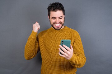 charming cheerful Young handsome Caucasian man wearing yellow sweater against gray wall holding in hands cell reading browsing