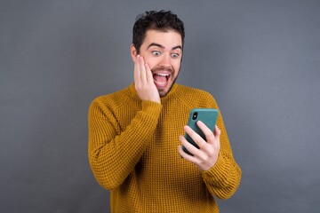 Young handsome Caucasian man wearing yellow sweater against gray wall holding in hands cell reading sms using new app 5g
