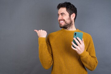 Young handsome Caucasian man wearing yellow sweater against gray wall using and texting with smartphone  pointing and showing with thumb up to the side with happy face smiling