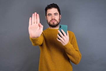 Young handsome Caucasian man wearing yellow sweater against gray wall using and texting with smartphone with open hand doing stop sign with serious and confident expression, defense gesture