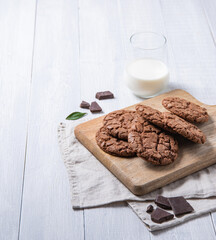 homemade chocolate cookies with chocolate chips on cutting board  and glass of milk  on a light wooden table