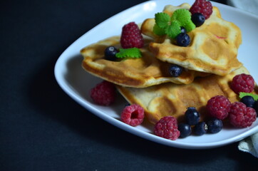 Homemade Belgium Waffles with fresh blueberries for breakfast. White plate, Viennese waffles with raspberries and mint. quick delicious breakfast on black background. top view copy space