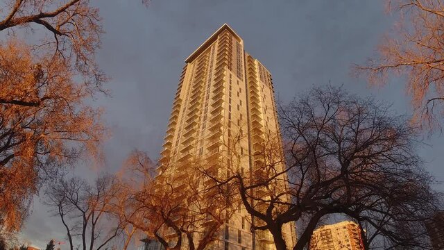 zoomout time lapse of clouds moving upwards and modern high tech futuristic apartment building with rooftop lounge glowing in gold natural lit sunset shinning on its corner view from the ground to top