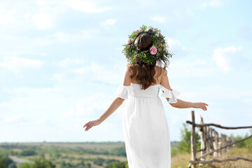 Young woman wearing wreath made of beautiful flowers outdoors on sunny day, back view