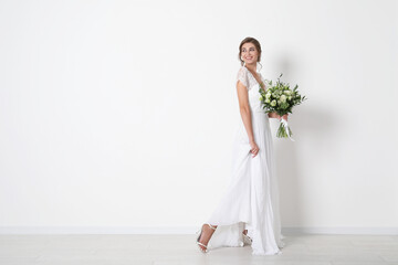 Young bride wearing wedding dress with beautiful bouquet near light wall. Space for text