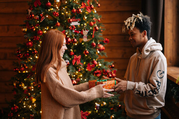 African American young man giving Christmas gift to Caucasian wife on background xmas tree with celebration lights at cozy living room with festive interior. Couple enjoying celebration.