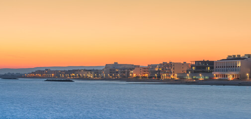 Vue d'une côte en bord de mer au coucher de soleil avec bâtiments et lumières du soir.