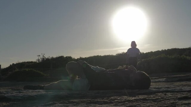 Silhouette Of Ambulance Officer With First Aid Bag Coming To A Man Lying On Clovelly Cliffs While Massaging Injured Knee - Paramedic - low angle