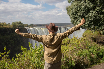 a man in a cowboy hat stands on a cliff against the background of Victoria Falls with his arms...