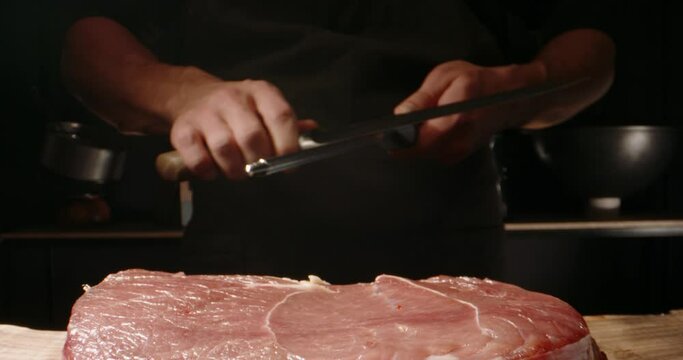 Chef sharpening his knife in front of raw piece of steak. Cooker preparing his tools for cutting meat for grilling on professional kitchen table 4k footage