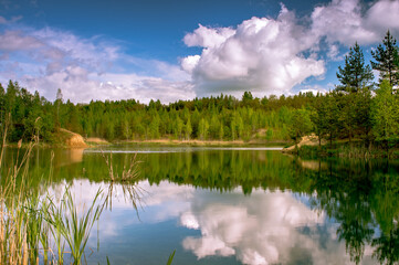 Lake with banks in bright spring green mixed forest with reflection of blue sky with clouds in clear water.