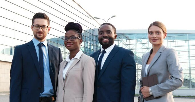Portrait of mixed-races young team of businessmen and businesswomen standing outdoor and smiling to camera on big modern business center background. Handsome sylish men and beautiful women.