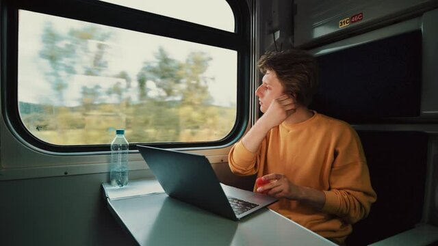 young man in wireless headphones rides in a train compartment and watches a video on a laptop with a serious face. Guy freelancer working on laptop at the table in the train.