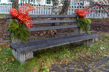 An old wooden park bench sits on green grass and among yellow leaves. There are two large red Christmas bows and garlands on the ends of the seat.  A white wooden picket fence is behind the chair.