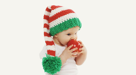 Portrait of baby in knitted gnome winter hat biting red apple over a white background