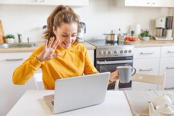 Smiling girl waving hand video calling family by webcam. Woman with laptop having virtual meeting chat video call conference sitting on kitchen at home. New normal social distance self isolation.