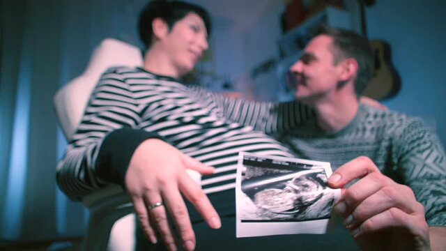Young couple posing. Pregnant woman is sitting in chair, next to her husband is holding an ultrasound photograph baby's fetus. Show the sympathy of young parents. Continuation of the human race.