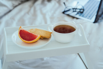 Selective focus of Breakfast in bed: white wooden tray with a cup of hot tea or coffee, a slice of grapefruit and cookies on the plate with a laptop and glasses in the background. Good morning concept
