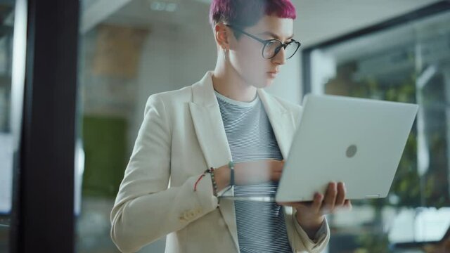 Modern Office: Portrait of Beautiful Creative Specialist with Short Pink Hair Standing, Holding Laptop Computer. Working on App Design, Data Analysis, Plan Strategy for Social Media Disruption 