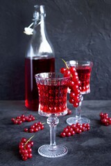 Red currant drinks in a beautiful glass goblets with bottle of currant drink on dark grey surface. Bunches of red currants on glasses and on the table