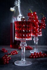 Glass with red currant drink in the foreground. A bottle and a glass with a drink in the background against a dark gray wall.