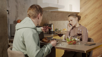 A young wife does not pay attention to her husband, talking on her smartphone during a festive family dinner with red wine. The man feels offended.