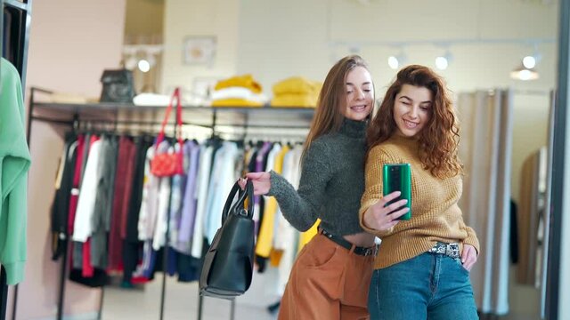 Two Stylish Girls Take a Selfie Photo on Smartphone or Phone in Front of a Mirror in a Clothing Store in a Shopping Mall. Young Happy Adult and Cheerful Shopaholic Women on Sale in a Boutique Indoors