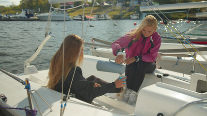 Young girls in sportswear relax on a yacht during a cruise in the port. They drink coffee from a thermos.