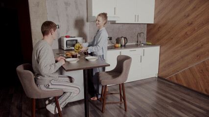 A woman serves a man at Breakfast. She brings him food on plates and puts cornflakes in the milk. The issue of gender inequality.