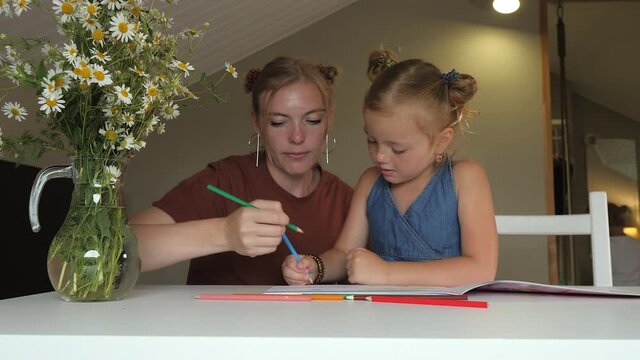 Mother and daughter draw together in a sketchbook with colored pencils sitting at the table at home. A large vase of flowers is standing next to it. Concept: homeschooling and quarantine.