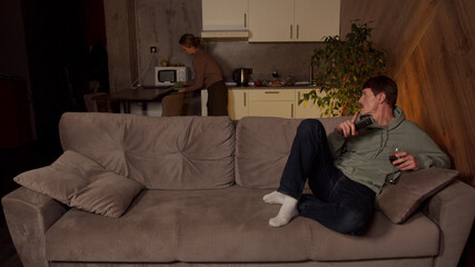 Obraz premium Husband with a remote control in his hands and a glass of wine watching TV, sitting on the couch. His wife is cleaning the kitchen.
