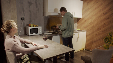 Cute blonde came to visit her friend, the guy makes a salad, she drinks wine in the kitchen