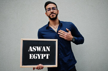 Arab man wear blue shirt and eyeglasses hold board with Aswan Egypt inscription. Largest cities in islamic world concept.