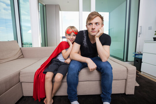 Portrait of smiling boy dressed in superhero costume sitting with sad father on sofa bed at home