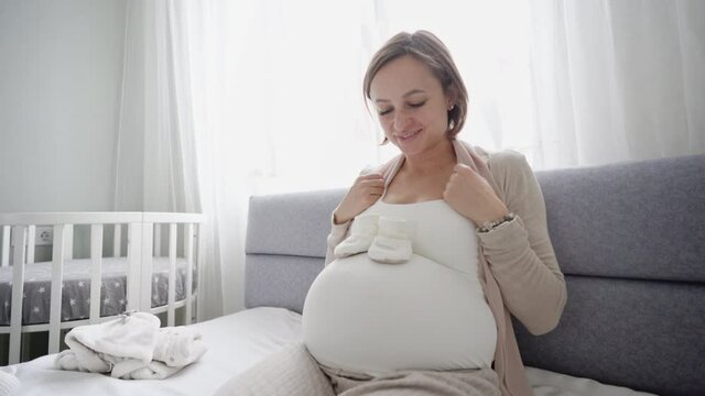 YounYoung pregnant woman with small baby shoes sitting on sofa at home. Expectant mother is preparing for childbirth. High quality 4k footage
