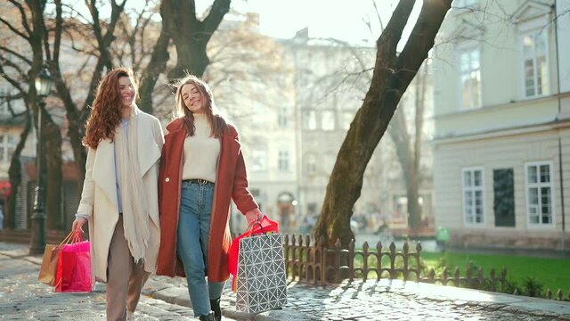 Two happy young women walking around the city after shopping on a city street in coats with gift bags in cold autumn winter or spring weather 2 cheerful girls go outside from a sale in a mall or store