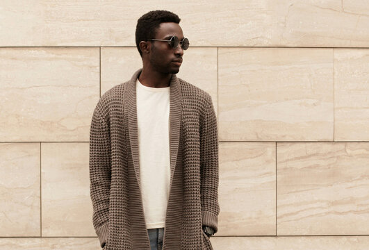 Stylish african man looking away wearing brown knitted cardigan, sunglasses on city street over brick wall background