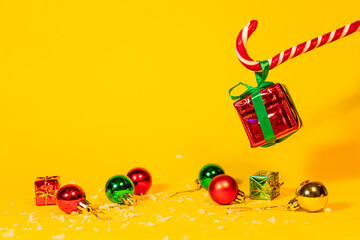 Candy cane lollipop holding a gift box with a Christmas present on a yellow background, Christmas sweets with new year decorations