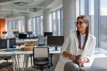 Obraz premium Portrait of a business smiling woman drinking coffee near the window in the office.