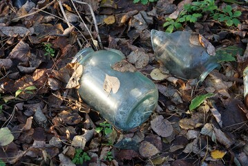 trash from one large gray dirty glass jar lies in brown fallen leaves in nature