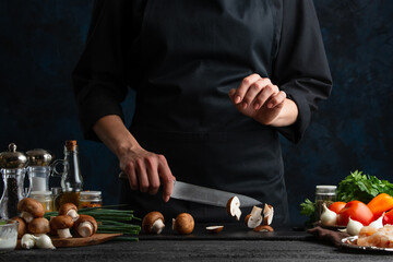 Professional chef cuts with knife mushrooms for preparing meat filling for pie. Ingredients for cooking on the background. Dark blue background space. Frozen motion. Cooking process.