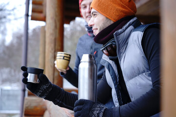 Two guys chat after skiing. They stand on the balcony of a wooden house in winter clothes, drink...