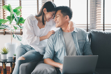 Portrait of happy Asian couple in love smiling and embracing together on sofa at home with laptop notebook